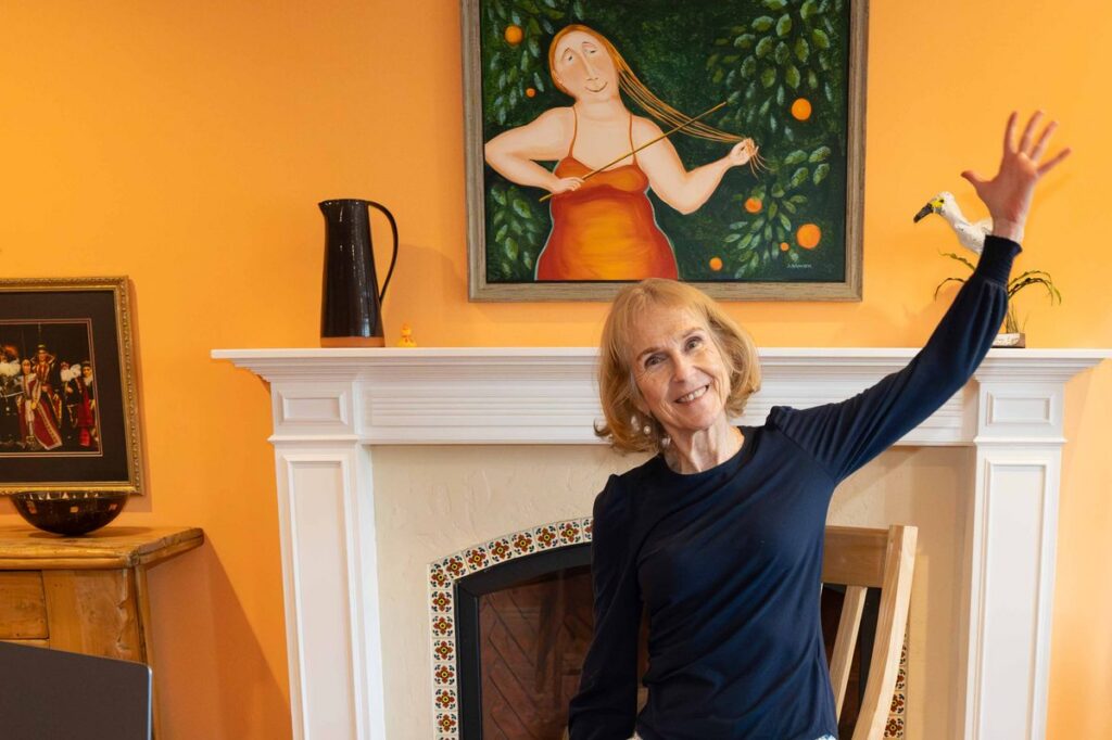 Patricia Becker teaching live online chair yoga, smiling with one arm raised in front of her laptop.
